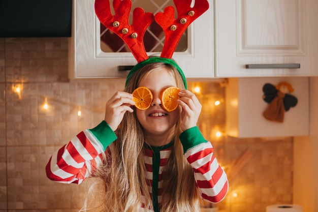 パジャマと枝角の小さな笑顔のかわいいブロンドの女の子は、彼女の目の近くに果物の半分を持っています。クリスマスツリーの近くのキッチンで子供。クリスマス
