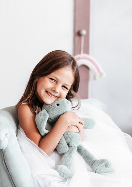 Little smiling cute girl in beautiful white dress is sitting with soft toy in her hands