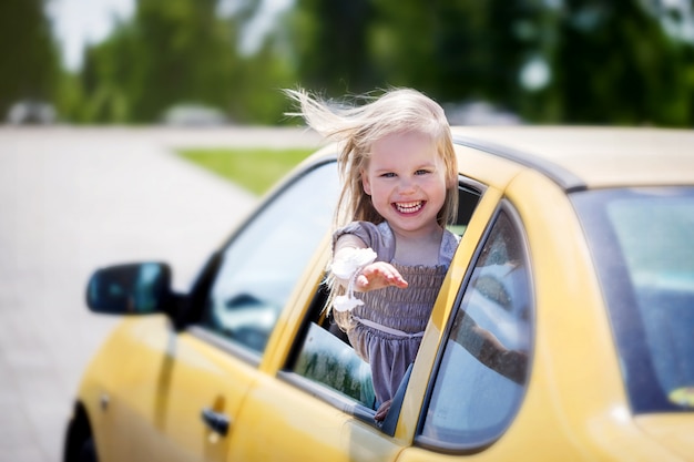 Little smiling girl is sticking her head out the car window and looking at camera