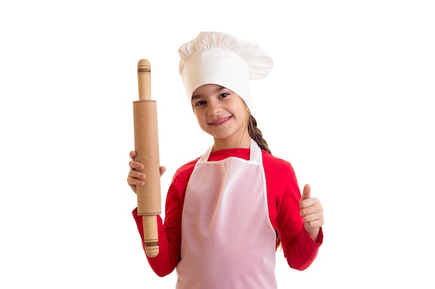 Little smiling girl in red shirt with white apron and hat holding rolling pin on white background