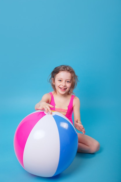 Little smiling girl in swimsuit with big inflatable ball on blue background