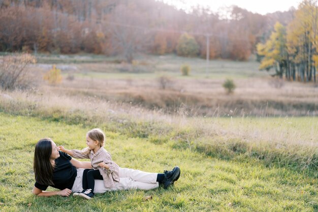 Little smiling girl touches her mother cheek lying on the green lawn sitting on her legs