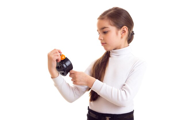 Little smiling girl with long brown ponytail in white shirt and black skirt holding orange clock