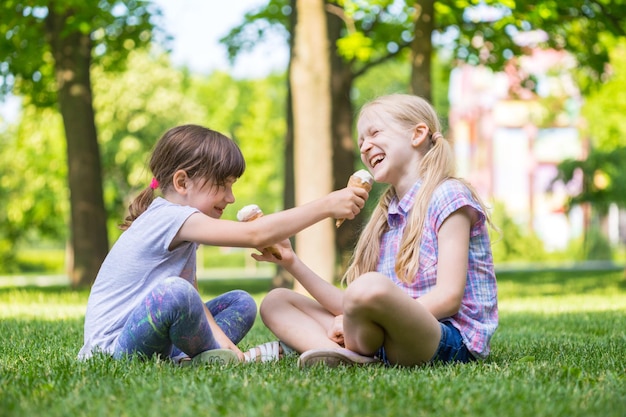 芝生の上に座ってアイスクリームを食べている小さな笑顔の女の子のガールフレンド。子供の休日