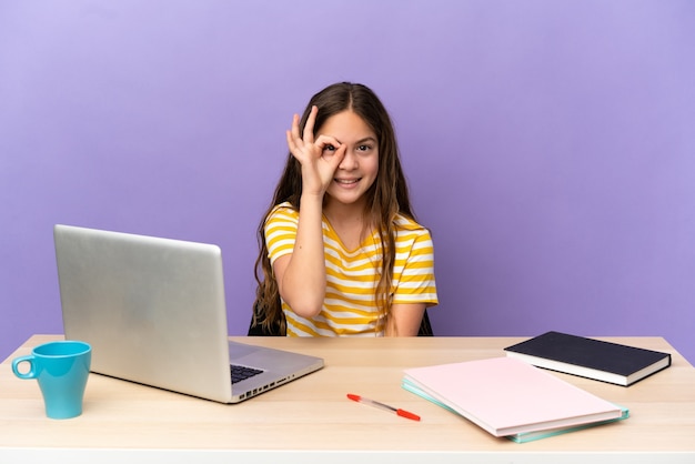 Маленькая девочка-студент на рабочем месте с ноутбуком, изолированным на фиолетовом фоне, показывая пальцами знак ОК