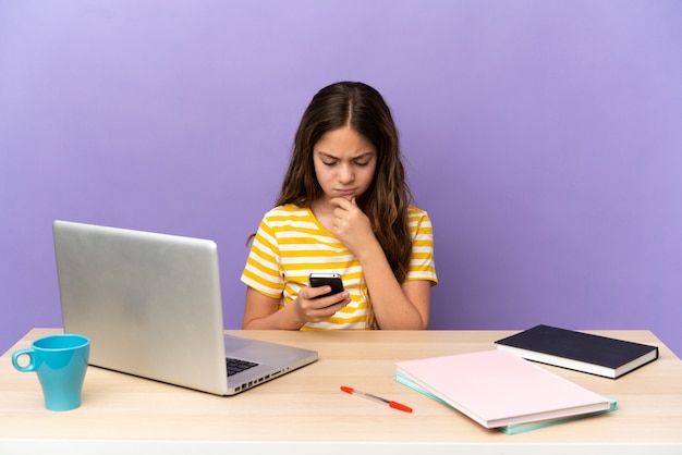 Little student girl in a workplace with a laptop isolated on purple background thinking and sending a message