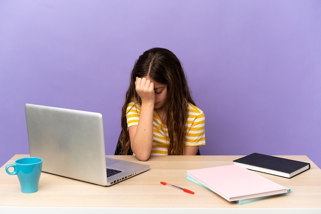 Little student girl in a workplace with a laptop isolated on purple wall with headache