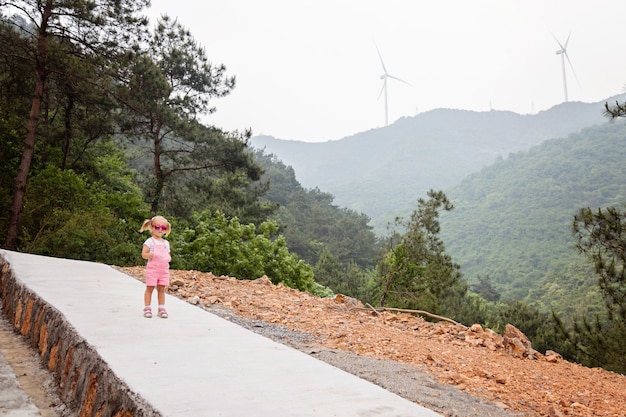 Piccola bambina alla moda con capelli biondi che cammina sulla montagna con i mulini a vento nel fondo. attività estive, vacanze locali durante la pandemia di covidi-19 di coronavirus