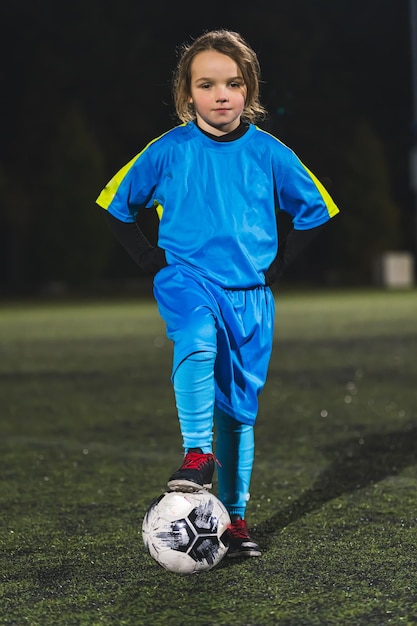 Little and super cute girl with a blue soccer uniform posing with a ball in a stadium at night