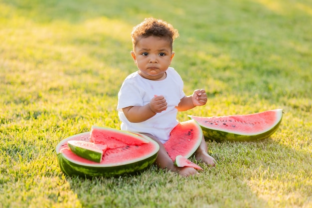 A little swarthy girl in white clothes eats a watermelon on a green lawn picnic in the park
