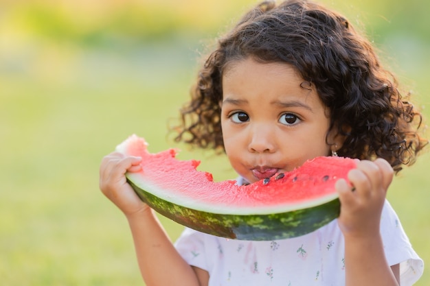 A little swarthy girl with curly hair in a pale pink dress eats a watermelon on the lawn