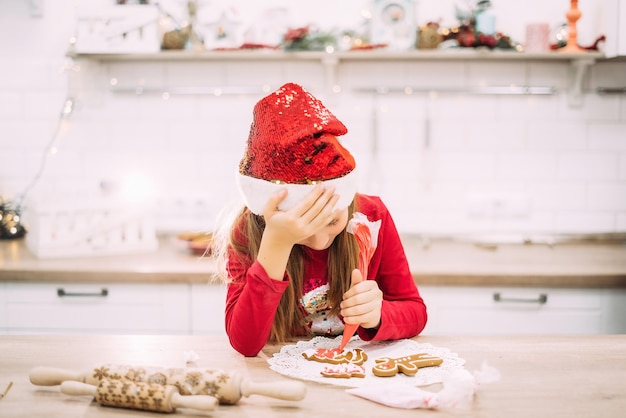 10代の少女は台所に座って頭を抱え、ジンジャーブレッドのクッキーにアイシングを塗っています。