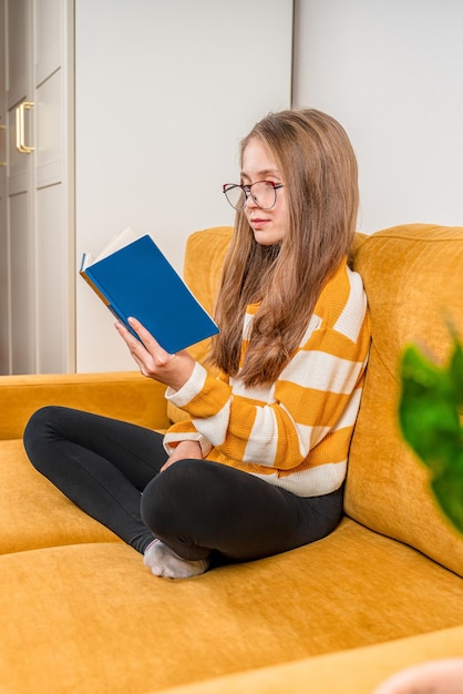 A little teenage girl with glasses is sitting on a yellow sofa at home and reading a book