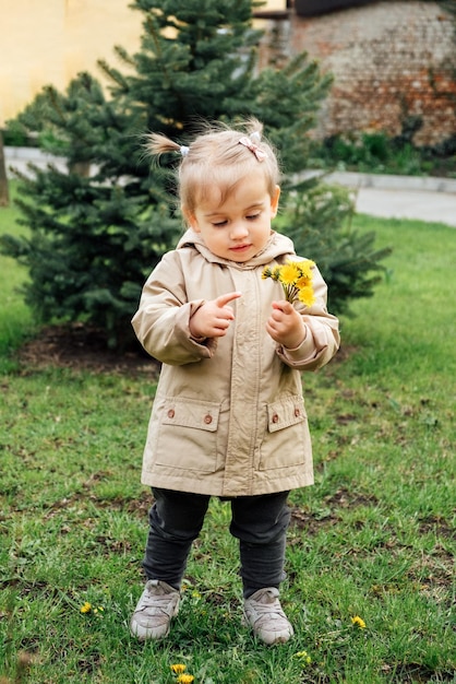 Bambina bambino piccolo in cappotto di trincea che raccoglie denti di leone gialli nel giardino di primavera bambina carina