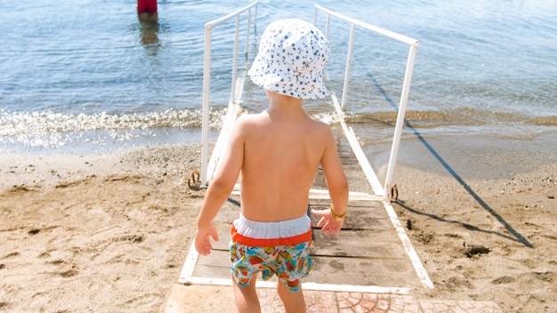 Piccolo ragazzo del bambino in cappello che cammina nell'acqua di mare sulla spiaggia.