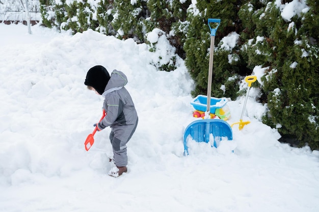 Little toddler boy in warm clothes with shovel cleans paths\
yard from snow kids winter games concept