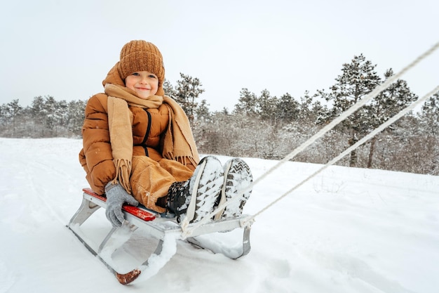 冬の服装で小さな幼児の子供が雪に覆われた公園でそりに座っています。