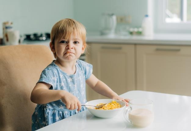 幼い女の子が朝にキッチンでミルクとコーンフレイクを食べる