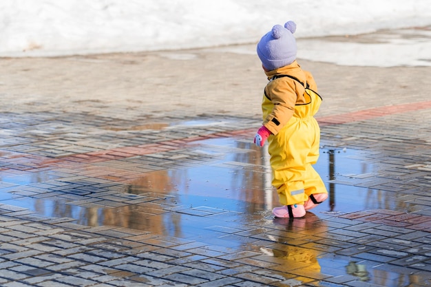 A little toddler girl runs through puddles in a yellow rubber jumpsuit