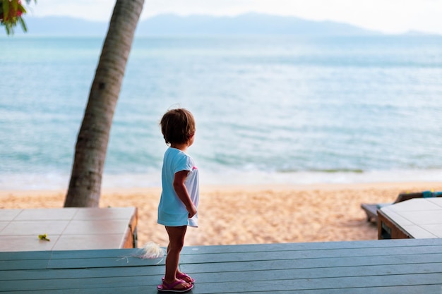 Little toddler girl in a white dress looks at the sea while standing on the wooden floor outside