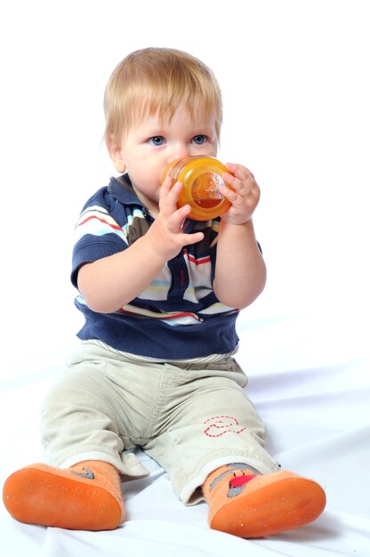 Il piccolo bambino si siede e beve l'acqua dalla bottiglia arancione