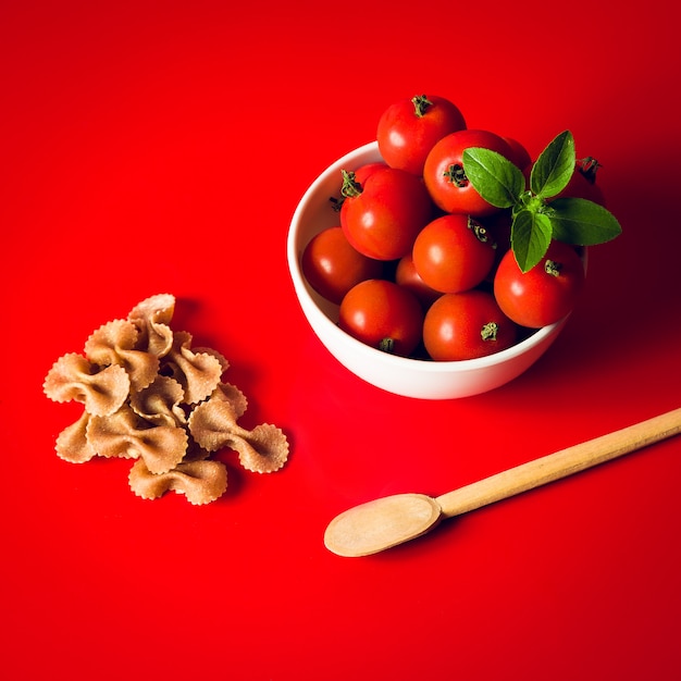 Маленькие помидоры в белой миске, с сырыми макаронами на красном фоне