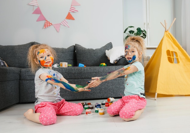 Little twin girls got dirty with paints for drawing sitting at home on the floor in the room