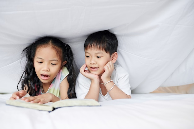 Little Vietnamese brother and sister lying under blanket and reading a book