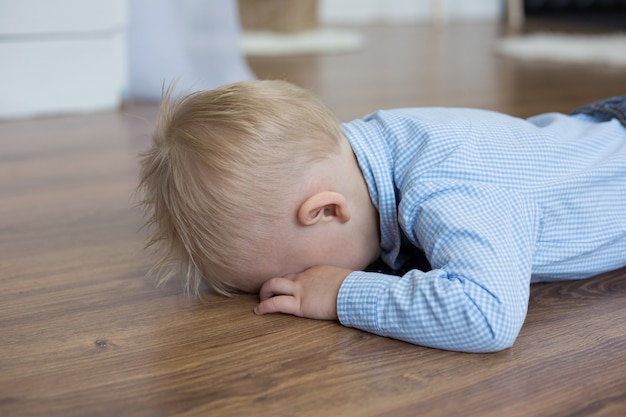 Un ragazzino bianco con una camicia blu si sdraia a faccia in giù sul pavimento e piange il concetto dei capricci dei bambini