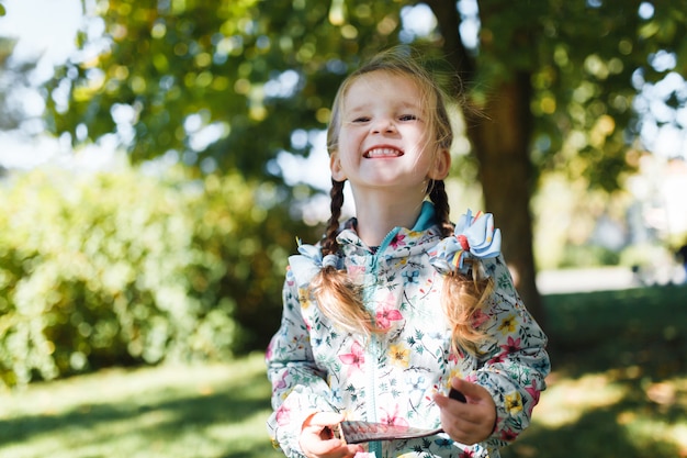 Little white happy girl with two pigtails in a multi-colored jacket looking at the camera and smiling on a warm autumn day