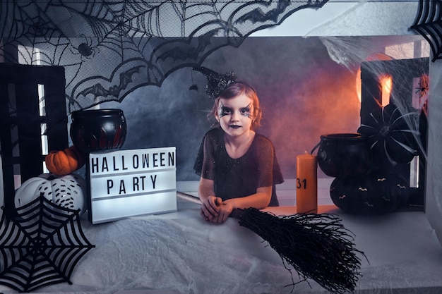 Little witch invite her friends for a party, she is sitting in well decorated area, there are spider web, pumpkins and broom.