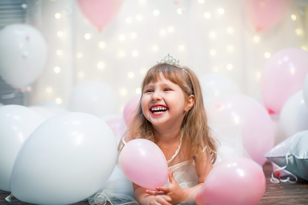 Little woman, in festive clothes and tiara, sits against balloons