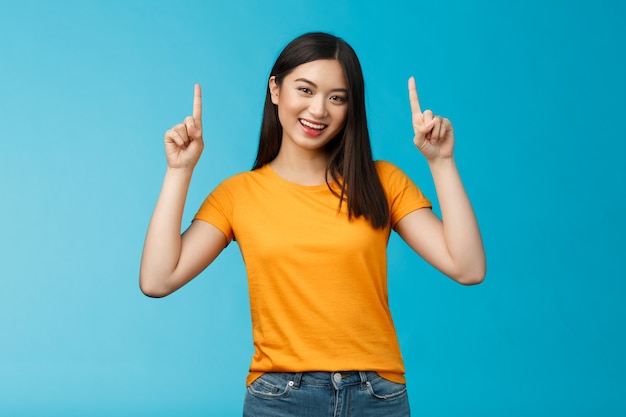 Lively asian girl dark short haircut, tilt head cute smile camera, raise hands and pointing index fingers up, introduce top promo, grinning, bragging new cool purchase, stand blue background.