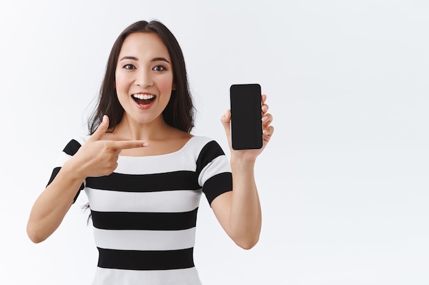 Lively, enthusiastic young asian woman excited introduce awesome new application, sharing favorite video or photo, describe her holiday to friend as pointing smartphone display, smiling joyfully