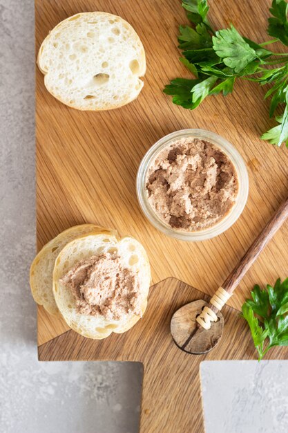 Liver pate in a glass jar with fresh bread and parsley on a wooden cutting board. 