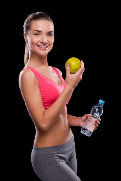 Living a healthy life. Happy young woman holding apple and bottle with water while standing against black background