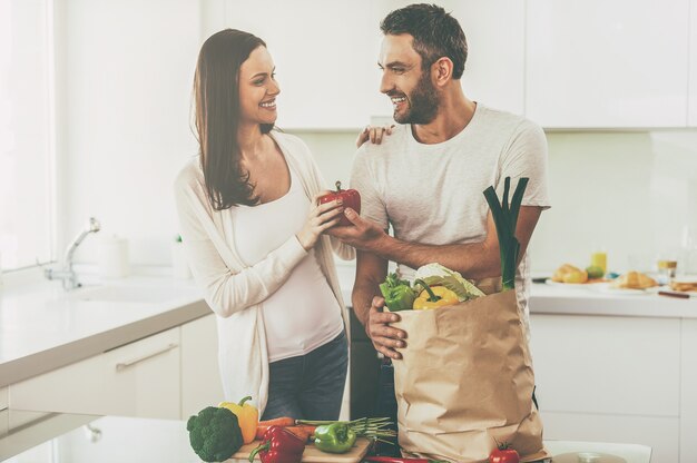 Living a healthy life together. Beautiful young couple unpacking shopping bag full of fresh vegetables and smiling while standing in the kitchen together