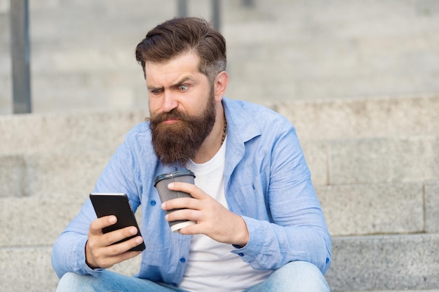 Vivere la vita online. social networks. comunicazione online della vita moderna. ragazzo con lo sfondo dello spazio urbano dello smartphone. bell'uomo barbuto rilassante cellulare e tazza di caffè. tecnologia moderna.