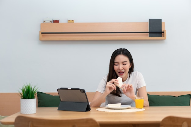 거실 개념은 직장 휴식 시간에 온라인 미디어를 시청하는 샌드위치와 오렌지 주스를 즐기는 여성 성인입니다.