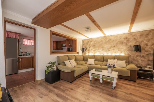 Living room furnished with a sofa with a beige chaise longue making a corner under a serving hatch leading to a kitchen furnished with wooden furniture