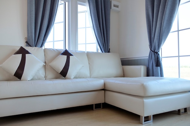 Living room inside the house with white leather sofa in the middle of a large window. And has a light gray curtain