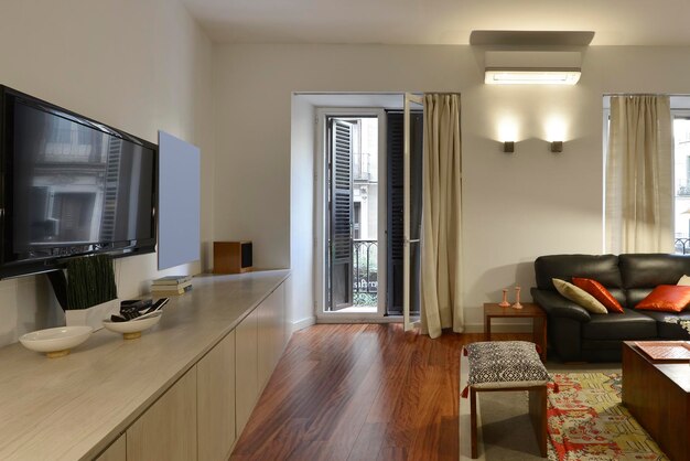 Living room in a studio apartment with a black leather sofa a walltowall wooden dresser and a flatpanel tv on the wall