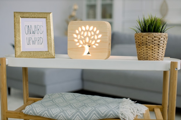 Photo living room with gray sofa and trendy coffee table with photo frame, decorative wooden lamp with tree picture, and green plant in wicker flower pot.