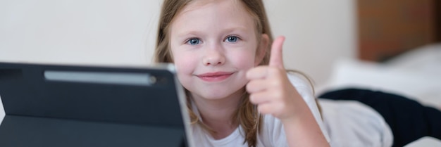 Photo llittle girl is lying on bed with tablet and holding thumbs up