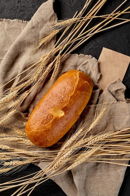 Loaf of freshly baked bread on cut of burlap with ears of wheat on black background