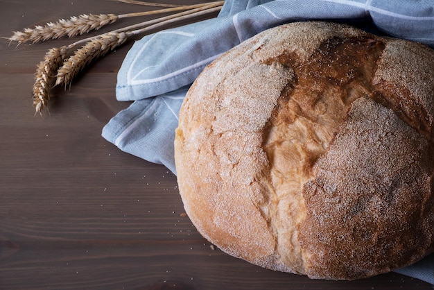 Loaf of freshly baked homemade artisan bread in linen napkin\
and spikelets with wheat grains on wooden surface selective\
focus