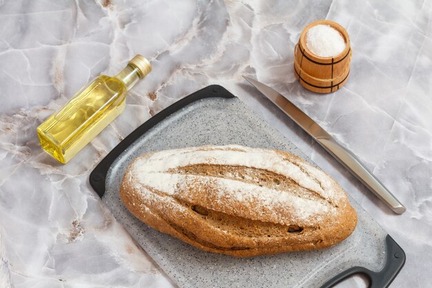 Loaf of rye bread on a cutting board, a bottle of sunflower oil, knife and a wooden salt cellar on the kitchen table