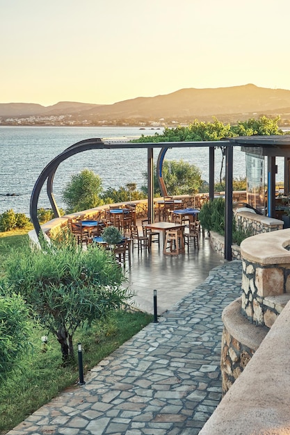 Local restaurant terrace with dining tables stone paved path and green trees against sea at sunset