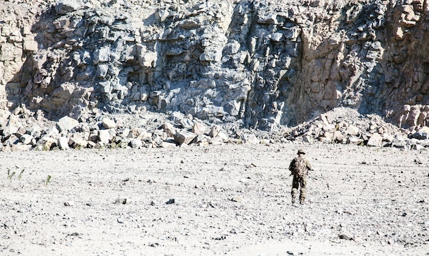 Colpo di posizione del soldato in uniformi da campo con il fucile che si muove nel deserto tra le rocce. vista posteriore