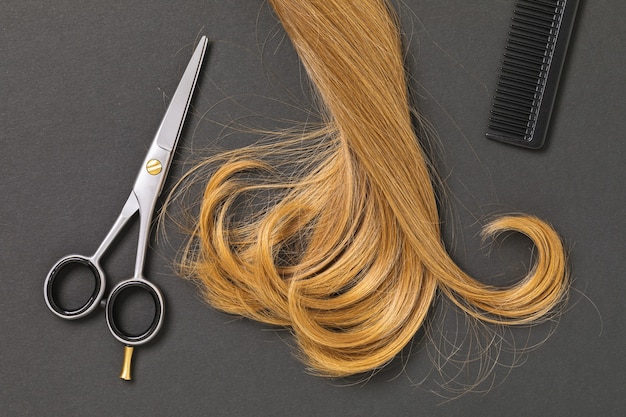 A lock of blond curly female hair, scissors and a comb on a dark background, top view